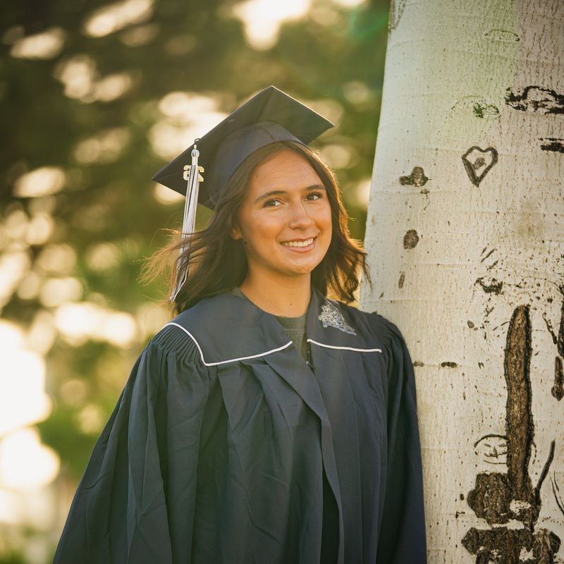 Graduate in cap and gown beside a white aspen trunk in warm backlight