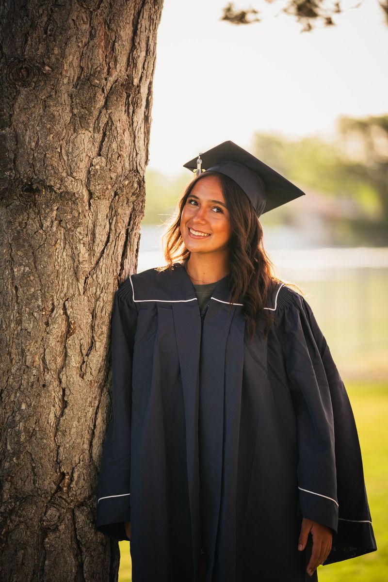 Graduate in cap and gown leaning against a tree trunk in evening backlight