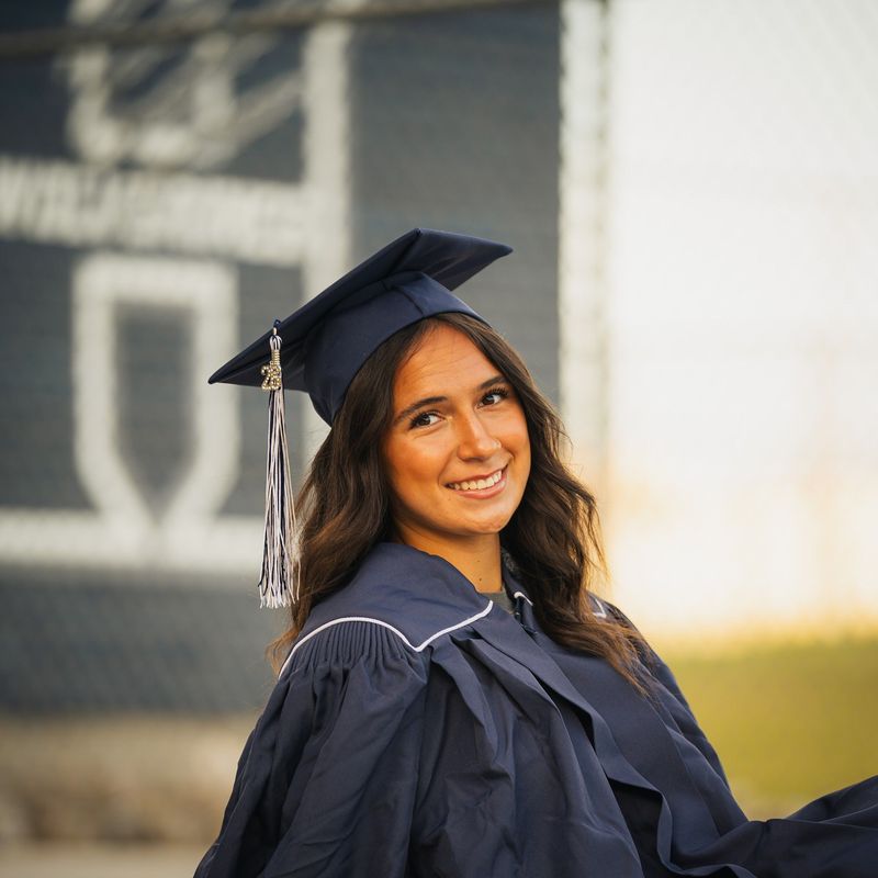 Close portrait of a graduate in cap and gown, school logo blurred behind