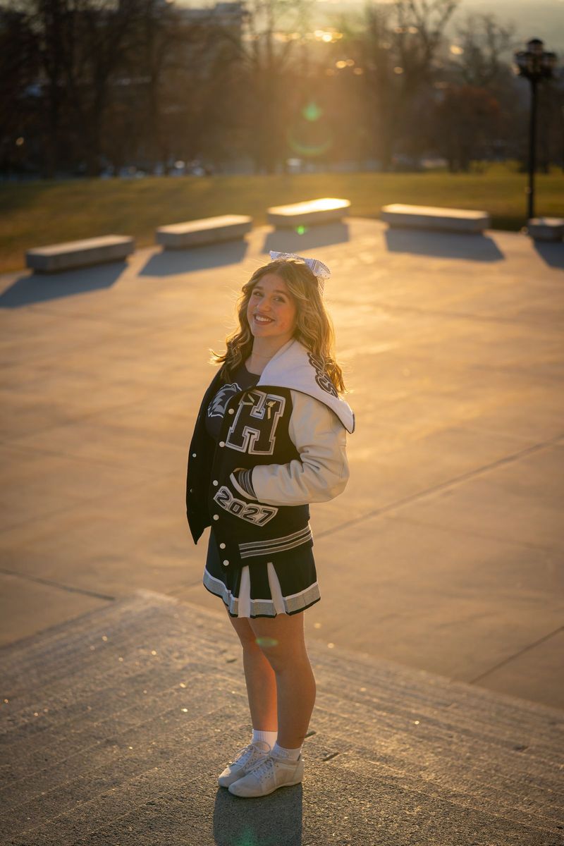 Cheerleader in a letterman jacket standing in a sunlit plaza with long golden shadows