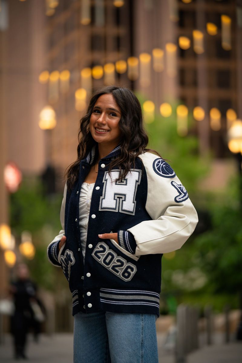 Senior in a letterman jacket, hands in pockets, warm city lights blurred behind