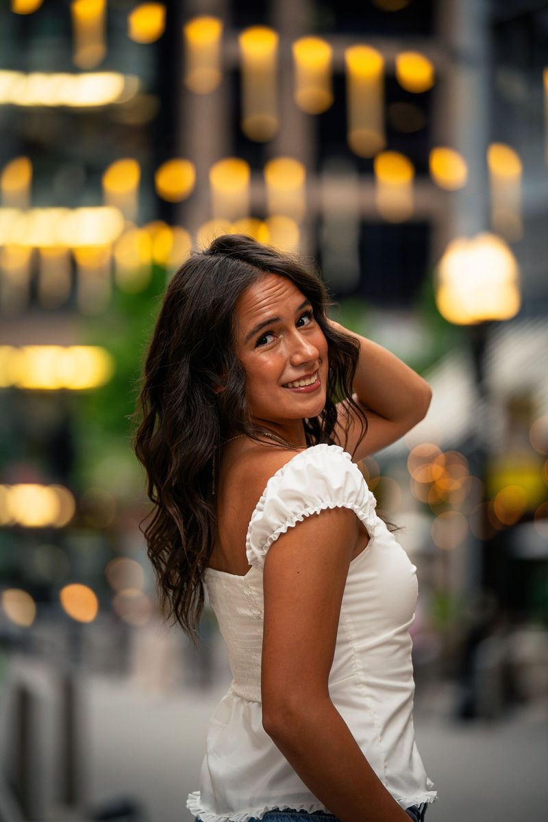 Senior in a white top with hand in hair, warm bokeh lights