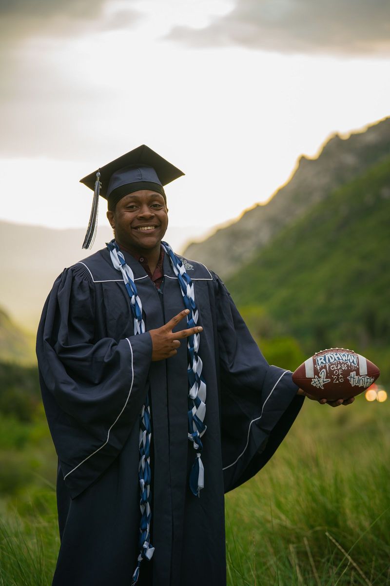 Graduate in cap and gown holding a football with a mountain ridge behind